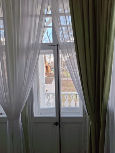 The view from inside a building in Borok, Russia, features a double-hung window partially obscured by olive-green and sheer white curtains. The upper window reveals a grassy yard with leafless trees and a glimpse of neighboring buildings, one with a decorative trim. The lower window offers a similar view, highlighting a rooftop and a building in the background. A decorative white trim with a swirl pattern is visible on one side of the upper window. The overall scene is a quiet, interior view looking out onto an early spring day in a residential area.