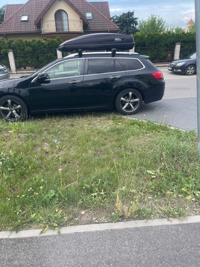 The image depicts a black station wagon with a black roof box parked on a street in Konstantynów Łódzki, Poland. The car is parked alongside a paved road and a grassy area with weeds and some small blue flowers. In the background, a residential building with a red-tiled roof and a balcony is visible, along with a fence and a row of green hedges. Another dark-colored car is parked further down the street. The sky is overcast.