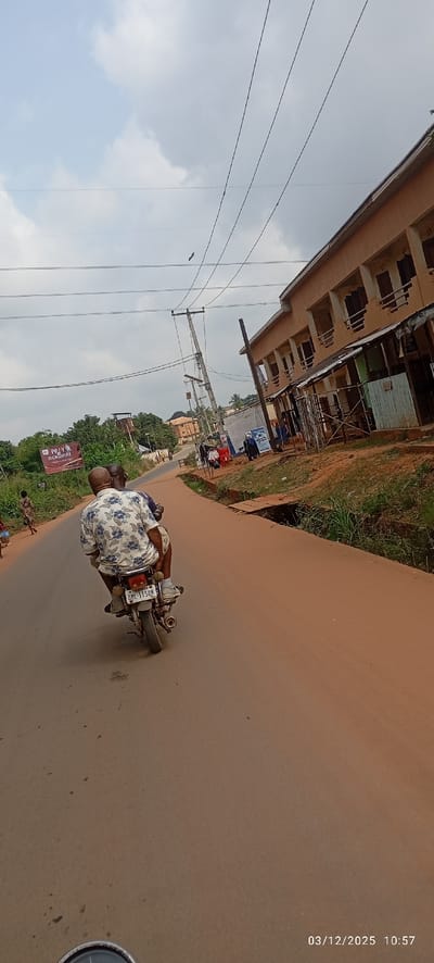 The image, taken on March 12, 2025, at 10:57, depicts a street scene in Abagana, Nigeria, captured from a low vantage point, likely from another vehicle or motorcycle.
In the foreground, two adult males are riding a light-colored motorcycle away from the viewer on a paved, reddish-brown asphalt road. The driver wears a light-colored, short-sleeved shirt with a blue floral pattern. The passenger, seated behind the driver, has a bald head and wears a darker shirt. The motorcycle's license plate, "UML 115QM," is visible.
To the right of the road, an open drainage ditch runs parallel, partially covered by concrete slabs in sections, with some green vegetation growing within it. Beyond the ditch, a multi-story building with an orange-brown facade and numerous open balcony/window areas is prominent. Below this building, closer to the road, are several smaller, single-story structures or stalls with corrugated metal roofing, indicative of commercial activity. Several individuals are visible near these structures. Multiple utility poles with overhead electrical wires extend along this side of the road.
To the left of the road, dense green vegetation, including trees and shrubs, lines the roadside. A large billboard with the text "PRAY OGBUNIKE" and an image (likely of Ogbunike Cave) is visible. Further down the curving road, additional residential or commercial buildings are discernible amidst the greenery. Another individual is seen walking on the left side of the road in the mid-ground.
The sky is overcast with significant cloud cover. A dark, bird-like object is visible in flight against the sky near the overhead utility lines in the upper central portion of the image.