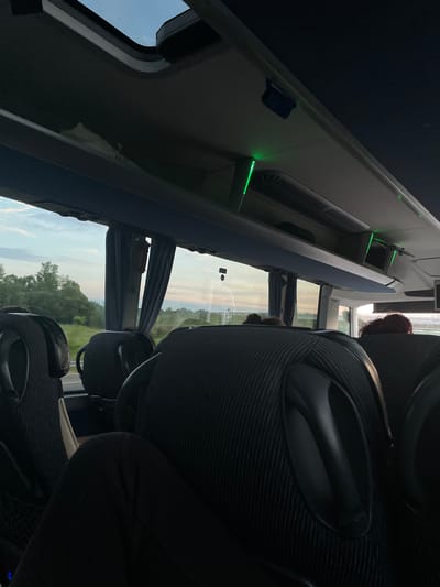 The image shows the interior of a bus, possibly a tour bus, with seats and luggage racks visible. The bus is moving; the view out the window suggests a rural landscape with trees and sky. The location is near Ub Municipality, Serbia.