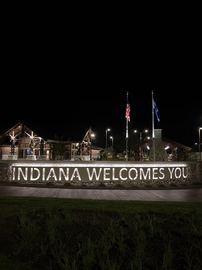This image captures the Indiana Welcome Center at night, located near New Harmony, Indiana, United States. The scene is illuminated by artificial lights, highlighting a large, illuminated sign that reads "INDIANA WELCOMES YOU." The sign is mounted on a stone wall and is backed by a textured pattern.
Behind the sign, the Welcome Center buildings are visible, constructed with log cabins and stone elements, suggesting a rustic architectural style. Two flagpoles stand prominently in front of the buildings, one displaying the American flag and the other a blue flag, possibly representing Indiana or another state entity. Several lampposts are also visible, providing additional lighting to the area. In the foreground, there is a patch of grass with some plants. The overall impression is one of a welcoming gateway to the state, observed at night. There are no people or animals visible in the frame.