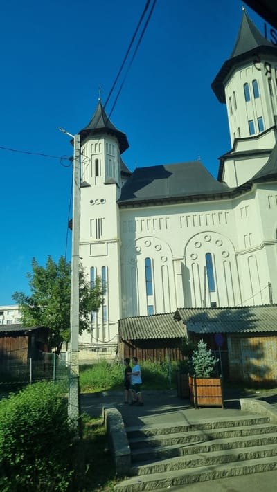 The image shows a white church with a dark roof and spire, located in Fălticeni, Romania. The church has several arched windows and decorative elements. In the foreground, two young males are standing on a sidewalk next to a set of stone stairs. One male is wearing a black t-shirt and shorts, and the other is wearing a white t-shirt and shorts. To the left of the church, there is a lush green tree and some smaller wooden structures with corrugated metal roofs. The sky is a clear, bright blue. The image is captured from a slightly elevated perspective, possibly from a vehicle.