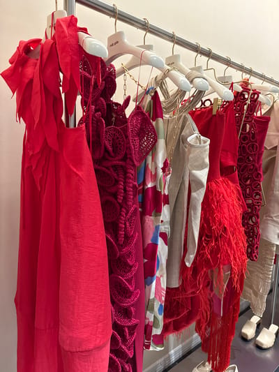 The image shows a clothing rack with various garments in shades of red, beige, and floral prints, against a light-colored wall. The garments are hanging on white hangers, and the rack itself is made of metal. From top to bottom, the garments appear to be: a beige garment, a red garment with circular cutouts, a red garment with fringe, a beige jacket, a silk garment with floral designs and red, crochet garments and a red garment with layered frills. The location appears to be inside a clothing store or studio in Sunrise, United States of America. There are no people visible.