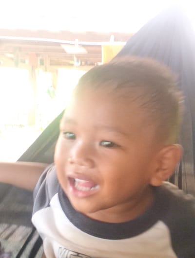 A close-up, slightly blurry, overexposed photograph shows a young child in an outdoor setting in Pompanua, Indonesia. The child, likely a toddler, is wearing a gray and white t-shirt. Their head is angled slightly to their right, and their mouth is open, revealing teeth. The background suggests a structure with wooden beams and possibly some hanging items, indicating an outdoor living space or covered area. A dark fabric, potentially a hammock or part of a shelter, is visible to the right and behind the child. The bright lighting suggests it is daytime, and the exposure makes it difficult to discern finer details of the surroundings or facial features.