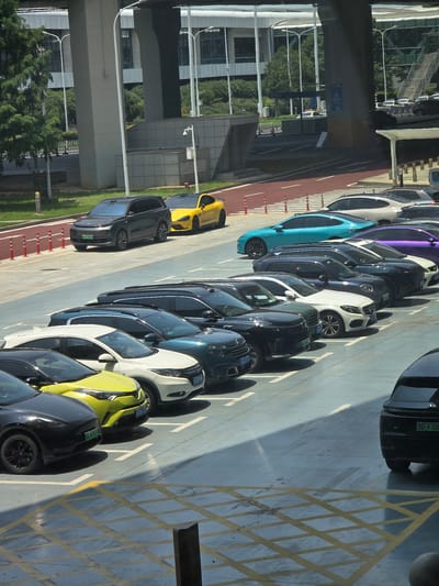A bright, outdoor, daytime photograph shows a multi-level parking lot in Wuhan, China, with numerous parked cars. The parking lot surface is concrete with yellow and white painted markings. In the background, a large, modern building with a glass facade is visible, along with trees and elevated roadways. Several vehicles are visible, including a dark gray SUV, a yellow sports car, a bright blue sedan, a purple sedan, a white SUV, a dark blue SUV, and other sedans and SUVs in various colors such as black and white. The cars are parked in designated spots, and some have license plates visible. The lighting suggests it is a sunny day, with strong shadows cast across the parking lot. There are no people visible in the image.