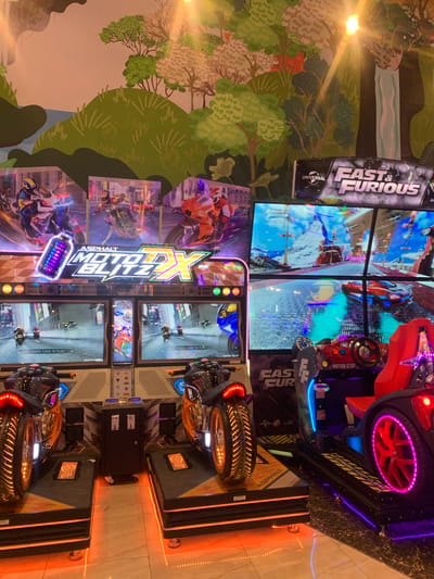 This media file captures a vibrant indoor arcade scene, featuring multiple modern racing game cabinets. The primary subjects are two types of elaborate arcade games: a "Fast & Furious" car racing simulator and several "Asphalt Moto Blitz DX" motorcycle racing simulators.

The "Fast & Furious" game, visible on the right, consists of a driver's seat, steering wheel, pedals, and a large curved screen displaying a high-speed race. Its platform features bright purple and blue LED lighting, and text like "MOTION STOP" and "NO STANDING KEEP CLEAR" is visible. A prominent vertical sign for "FAST & FURIOUS" with the "UNIVERSAL" logo stands next to it.

Arranged on the lower left and middle are multiple "Asphalt Moto Blitz DX" motorcycle racing games. Each features a motorcycle-shaped controller with handlebars, a seat, and dedicated screens showing gameplay. These games also incorporate bright orange and blue LED lighting, especially around the wheels and bases. Screens on these machines display "SWIPE CARD TO PLAY," indicating they are ready for use, though no one is actively playing in the image.

The background features a colorful wall mural depicting a stylized forest with green trees and a light blue waterfall. The overall setting appears to be a brightly lit entertainment center, likely in Samarinda, Indonesia, given the context. The artificial lighting from the games and overhead fixtures creates reflections on the polished floor and game surfaces. The image is taken at an angle, and the scene is illuminated by the glowing screens and LED strips of the arcade machines.