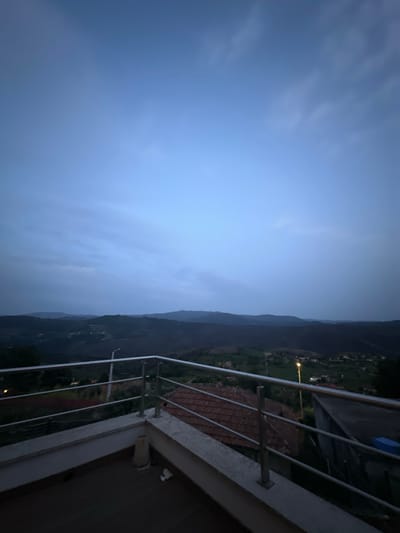View from a high vantage point, likely a balcony, in Celorico de Basto, Portugal. The sky is overcast and blue, suggesting dusk or dawn. The land is a valley with rolling hills and mountains, and the houses and buildings are visible in the background. The balcony railing is white metal and runs along the right side of the image. The balcony floor is dark brown.
