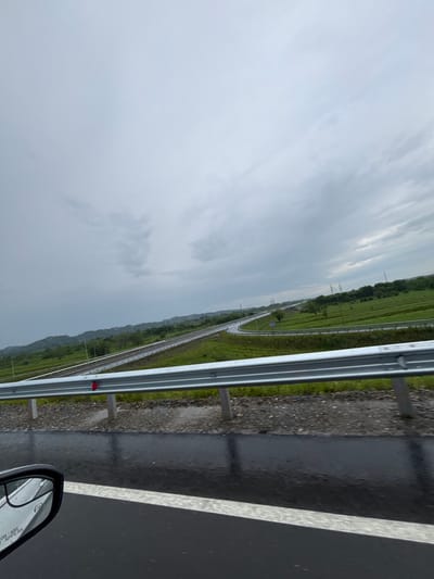 The image depicts a highway scene on a cloudy day, with a focus on the road and its surrounding landscape. A silver guardrail runs horizontally across the foreground, with metal posts supporting it. The road itself appears wet, suggesting recent rain, and a white line is visible marking a lane. To the left, a car's side mirror is partially visible, reflecting the overcast sky and showing text that reads "OBJECTS IN MIRROR ARE CLOSER THAN THEY APPEAR." The background features a winding highway that disappears into the distance, flanked by green fields and rolling hills. Several utility poles are scattered throughout the landscape. The sky is filled with heavy, grey clouds, indicating a somber or overcast atmosphere. The scene appears to be an open, rural or semi-rural area. There are no people visible in the image. The location is identified as Lesi Municipality, Georgia.