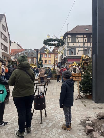 The image displays an outdoor Christmas market scene in Sélestat, France, under a uniformly overcast sky. The ground is paved with irregular light-colored cobblestones. In the foreground, a person wearing a dark olive-green hooded jacket, dark pants, and two-tone green and black boots stands with their back to the camera, carrying a light green tote bag. Adjacent to this individual is a black metal brazier, approximately 0.7 meters tall, containing glowing embers and resting on three legs. To the right, a young boy, also facing away from the camera, is clad in a dark blue puffer jacket, gray pants, a dark knit cap, and light brown boots. Immediately to the boy's right, loose, rough-hewn stones lie on an area of disturbed ground.

The midground features approximately 20-30 individuals, dressed in various winter coats, hats, and scarves, moving and congregating within the market. A vertical wooden sign is visible, displaying "Marché de Noël" and "Weihnachts Dürfle." Several small, decorated Christmas trees with gold baubles are present, signaling market stalls. A prominent central feature is a tall pole structure supporting a large circular evergreen wreath adorned with gold baubles and stars. Two illuminated, heart-shaped lanterns are mounted at the top of this structure. In the background, traditional European buildings line the street, notably a half-timbered building on the right with light pink infill and dark window shutters. Other structures are light-colored stucco. Two distant architectural features, topped with heart-shaped lanterns, are visible above the central wreath. Market stalls, some illuminated with string lights, extend into the background. A wooden fence encloses a seating area with picnic tables and benches to the left midground, where several people are seated.