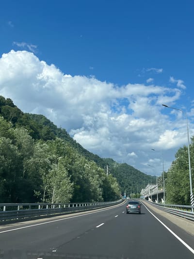 An eye-level, forward-facing shot captures a roadway in Kepsha, Russia, under a bright blue sky dotted with white clouds. The highway has two lanes in each direction, separated by a median, and a metal guardrail lines the outer edges. Green trees and densely wooded mountains flank the left side of the road, while a bridge and more trees are visible on the right. A silver car is in the right lane of the oncoming traffic, and other vehicles are further ahead on the road. Streetlights are positioned along the highway.