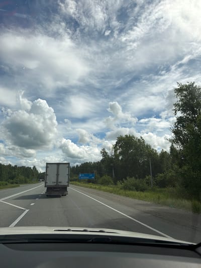 The image depicts a view from inside a vehicle, looking out at a two-lane asphalt road in a rural setting. A white truck with a closed trailer is driving in front of the vehicle. A blue road sign is visible on the right side of the road, indicating "УФА 446" and "ПИТОМНИК 1" with directional arrows. The road is flanked by dense green trees and vegetation on both sides, suggesting a natural landscape. The sky is partially cloudy with a mix of white cumulus clouds and thinner cirrus clouds against a blue sky. The overall scene suggests a journey on a highway in Russia, potentially near Poletaevo 1th based on the provided information, on a day with variable weather. There are no people visible in the image.