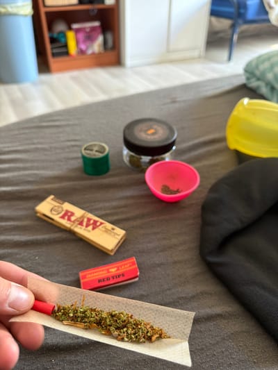 The image shows a close-up of a person's hand rolling a cigarette on a grey fabric surface. The cigarette is partially rolled and filled with a green, herbaceous substance and appears to have a red filter tip. Scattered around are various items associated with rolling cigarettes, including a pack of "RAW" rolling papers, a small red box labeled "DIAMOND LEAF RED TIPS," a glass jar with a black lid containing a similar green substance, a small green cylinder, a bright pink bowl, and a yellow container with a lid. In the background, a blurry wooden shelf unit is visible, with some items on its shelves, and a blue chair can be seen. The overall scene is indoors, likely in a residential setting. The image does not contain any information that can be used to determine its location within the city of Boxtel, Netherlands.
