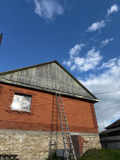 In the city of Bolshoy Turysh, Russia, an exterior shot captures a brick building under a bright blue sky with scattered white clouds. A wooden ladder leans against the red brick facade of the building, extending towards the grey, weathered wooden attic. A single window, reflecting the sky and clouds, is visible on the upper brick level, above a foundation made of rough, light-colored stones. A brown metal door is situated lower on the brick wall. To the right, a corrugated metal fence encloses a darker, rustic structure, partially obscured by green grass. The scene suggests a rural or semi-rural setting, possibly a home or outbuilding, with signs of maintenance or repair evident from the ladder's presence.