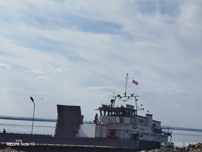 The image depicts a vessel identified by the name "ОТКОС" on its superstructure, flying the Russian flag. The vessel is situated on a body of water, likely a river, given the distant shoreline visible. In the foreground, there is a section of a dock or pier, with a lamppost and two people visible on it. The sky is partly cloudy, with a light blue hue. A timestamp "07.07.2026 16:59" is present in the bottom right corner, along with the text "REDMI Note 13" in the bottom left. The scene is consistent with a location in Nizhny Bestyakh, Russia.