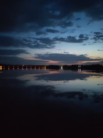 The image depicts a body of water at twilight in Ermolino, Russia. The sky is filled with dark clouds, tinged with blue and orange hues near the horizon. Reflected in the water are the clouds and faint lights from across the water. These lights are likely from structures or streetlights. A row of red lights is seen in the distance, also reflected in the water. The foreground of the image is dark, suggesting the perspective is from the water's edge or a low vantage point. No people or identifiable subjects are visible. The overall environment suggests a calm, possibly rural or suburban, setting.