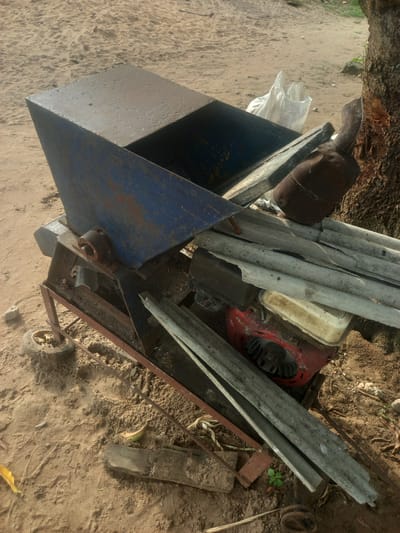 In Ikoga, Nigeria, a weathered blue hopper machine, likely used for processing agricultural produce, rests on sandy ground near the base of a tree. The machine's metal surfaces are rusted and show signs of wear. A small, worn wheel is attached to its frame, suggesting it is portable. Several lengths of corrugated roofing sheets are piled on top of the machine and lean against its side. Beneath these sheets, a red engine with a white fuel tank is visible. A white plastic bag lies next to the hopper, and various pieces of debris, including wood scraps and dried leaves, are scattered around the machine. The scene is illuminated by natural daylight.