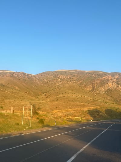 The image shows a road in Arpi, Armenia, with a clear blue sky overhead. The road is paved and has white lane markings. To the left of the road, there is a grassy embankment with a white rectangular frame structure. Beyond the embankment, rolling hills covered in dry grass and scattered rocks rise towards the horizon. A transmission tower is visible on the right side of the image, situated on the hillside. The overall scene depicts a rural, arid landscape under a bright sky.