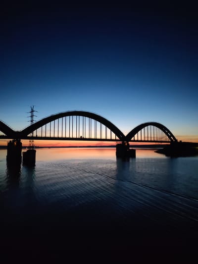 The image captures a panoramic view of a large, twin-arch through arch bridge silhouetted against a twilight sky over a body of water in the city of Zarya, Russia.
The sky exhibits a pronounced gradient, transitioning from a dark navy blue in the upper third to a medium, deep blue through the middle, and finally to a vibrant band of orange, pink, and red hues along the horizon line, indicating either sunrise or sunset. A faint white speck, possibly a star, is visible in the upper right quadrant of the sky.
The bridge, the primary subject, is rendered as a dark, intricate silhouette. It features two prominent, symmetrical arched spans, each supported by numerous vertical hangers extending downwards to the bridge deck. The bridge structure is anchored by two large, dark, cylindrical piers submerged in the water near the center-left. A small, dark triangular sign with an indistinct symbol is visible on the bridge deck within the right arch.
To the far left of the bridge, a tall, metallic lattice-work power transmission pylon stands silhouetted against the lighter blue portion of the sky. The right bank of the water body appears as a dark, indistinct landmass, with a small, dark structure barely discernible on its edge.
The water surface, likely a river or lake, is dark, reflecting the deep blues of the sky above and the vivid orange and pink glow from the horizon, particularly beneath the bridge. Subtle ripples and wave patterns are visible across the water, especially in the foreground and around the bridge piers. No discernible human figures, vehicles, or vessels are present in the scene.