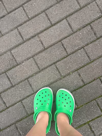 The image shows a pair of bright green Crocs shoes on a person's feet, standing on a grey brick pathway. The pathway is laid in a herringbone pattern and has some patches of moss growing in the gaps between the bricks. The perspective is from directly above the feet, looking down. The scene appears to be outdoors, possibly a park or a pedestrian area within a city. Without further information or identifying landmarks, it is not possible to determine the specific location within Rzeszów, Poland.