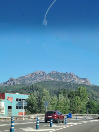 The image captures a scenic view of the mountains of Olesa de Montserrat, Spain, under a clear blue sky. In the foreground, a red SUV is driving on a road. The road is marked with white lines and bordered by blue and white striped traffic posts. To the left of the road, there is a turquoise building with red trim and the word "CMP" in orange lettering. The building has several windows and an orange door. Behind the building and the road, a dense forest covers the lower slopes of the mountains. The mountains themselves are rocky and rugged, with a distinctive jagged skyline. The overall impression is one of a beautiful, natural landscape juxtaposed with a touch of human infrastructure.