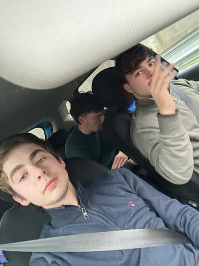 Three young men are visible in a car, seemingly in the backseat. The man at the top is wearing a gray sweatshirt. The man in the middle appears to be wearing a green shirt. The man in the bottom is wearing a blue jacket and seatbelt. All three appear to be looking towards the camera. The interior of the car is visible, including the seats and headrests.
