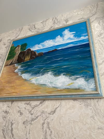 A framed oil painting of a beach scene is hung on a wall with marble-patterned wallpaper. The painting depicts a sandy beach with waves crashing on the shore. In the background, there are rocky cliffs and a blue sky with white clouds. The frame is ornate and appears to be light blue and silver. The scene is indoors, with the painting lit by an overhead light source, casting a slight shadow on the wall. The image does not contain any people or specific identifiable landmarks of Gaverdovskii, Russia.