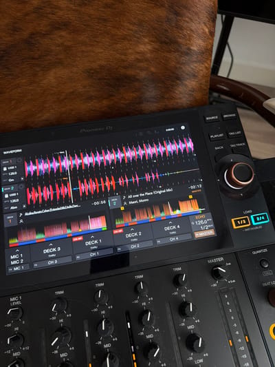 The image shows a close-up view of a Pioneer DJ controller and a portion of a mixer. The DJ controller's screen displays waveforms for two music tracks, labeled "Deck 1" and "Deck 2," along with track information. The mixer features several knobs and buttons for audio control. The device is positioned on a surface covered by a textured material, likely an animal hide, with a brown and black brindle pattern. The background is blurred, but suggests an indoor setting with some lighting. No people are visible in the image. There is no information to suggest this scene is located in Haarlem, Netherlands.