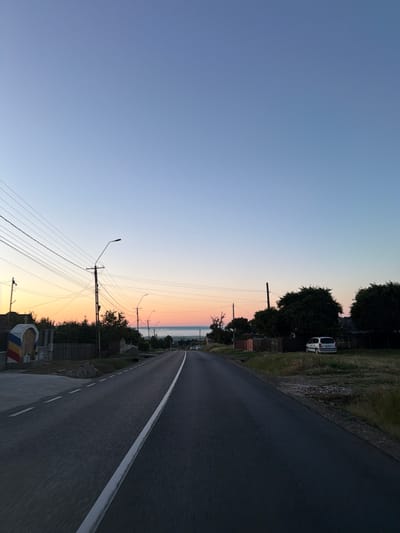 The image depicts a road in the outskirts of Pașcani, Romania, at dawn or dusk. A paved road with a white central line stretches into the distance, curving slightly to the right. Overhead power lines crisscross the sky, supported by utility poles. On the left side of the road, there is a white structure adorned with red, yellow, and blue bands, resembling a monument or memorial, adjacent to a wooden fence. A few houses and trees are visible along the roadside. On the right side, a white car is parked on a grassy area next to a fence, with more trees and houses further back. The sky transitions from a light blue overhead to warmer hues of orange and pink near the horizon, indicating the presence of the sun just below or rising above the landscape. The overall scene conveys a sense of rural tranquility.