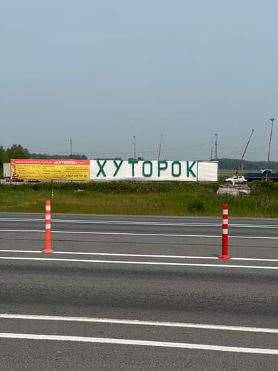 A wide shot captures a roadside advertisement in Kargat, Russia. The sign, predominantly white with large green lettering, proclaims "ХУТОРОК" (Khutorok), with smaller text in red indicating "1281 км" (1281 km). To the left of the large green lettering, a smaller red banner displays text including "ДОБРО ПОЖАЛОВАТЬ К НАМ НА" (Welcome to our) and a list of amenities such as "Кухня. Русская и Восточная. Баня на дровах. Магазин. Гостиница. Санитарный комплекс. Охраняемая парковка. Сто. Беседки." (Kitchen. Russian and Eastern. Wood-fired bathhouse. Shop. Hotel. Sanitary complex. Guarded parking. Service station. Gazebos.) Below this list, a phone number is visible: "8 913 986-55-32". The sign is positioned in a rural setting with grassy fields and trees in the background. A white vehicle and a blue truck are parked behind the sign. In the foreground, a paved road with white lane markings is visible, with two orange traffic cones with white stripes placed on the shoulder. The sky is overcast and pale blue.