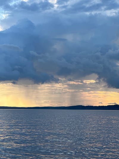 The image shows a wide expanse of water under a dramatic sky. Dark, heavy clouds dominate the upper portion of the frame, with lighter shafts of sunlight breaking through near the horizon, creating a streaky, diffused light effect. The sun appears to be low in the sky, casting a warm, golden glow. A dark, tree-lined landmass is visible as a distant silhouette across the water, marking the shoreline. The water itself is calm, with a rippled surface reflecting the colors of the sky. The scene is serene and atmospheric, suggesting either dawn or dusk. There are no people or man-made structures clearly visible in the image. The location is identified as Shul'dikha, Russia, implying this is a natural landscape scene within that region.