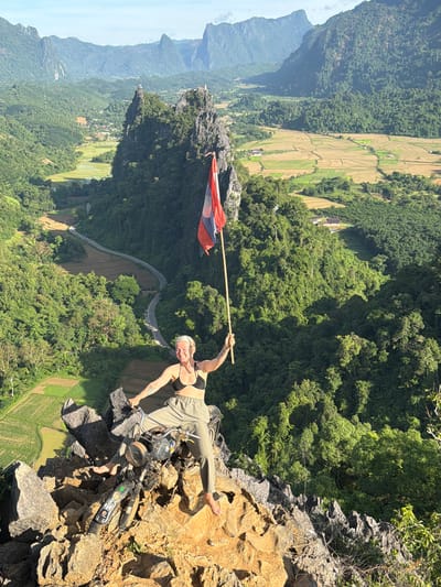 A woman stands on a rocky precipice in the city of Naka, Laos, holding a flag aloft. She is wearing a black bikini top and olive-green pants, with her left leg extended forward and her right leg bent. Her arms are raised, and she is looking towards the camera with a smile. A motorcycle is parked behind her, partially obscured by her legs. The background is a lush, green landscape of rolling hills and rice paddies, with a winding road visible in the distance. The sky is a clear blue with a few wispy clouds. The flag she holds is red, white, and blue, with a depiction of a mythical creature in the center.