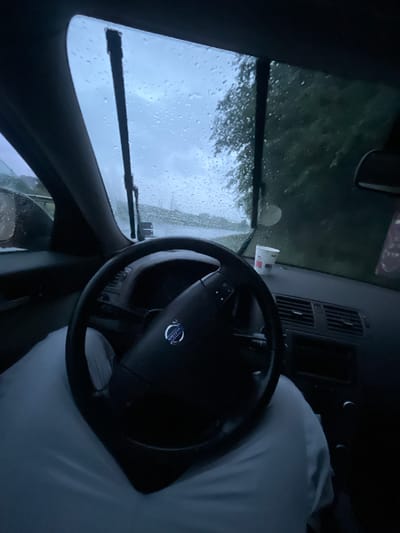 The image depicts the interior of a vehicle, specifically focusing on the driver's perspective. Rain is visible on the windshield, with wipers actively clearing the water. The steering wheel is prominently featured, displaying the emblem of what appears to be a Volvo. An object resembling a white coffee cup is placed on the dashboard. The presence of a white, shapeless form near the steering wheel suggests a person covered by a blanket. The location is ambiguous, as there is not sufficient detail to identify the location as Damianitsa, Bulgaria.