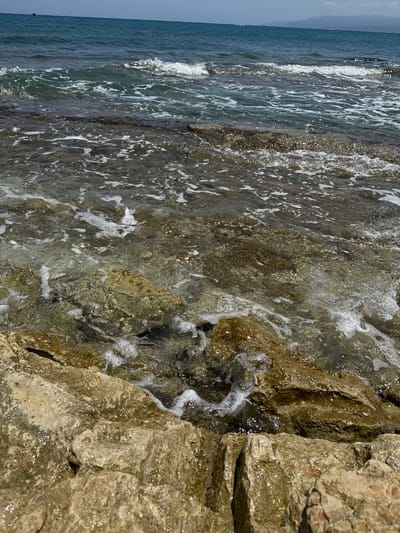 The image shows a rocky coastline with clear water gently lapping against the shore. The foreground is dominated by large, rough, light brown rocks, some of which are wet and glistening. The water is shallow, allowing the submerged rocks and seabed to be visible. Small waves are breaking, creating white foam and ripples on the surface. In the middle ground, the water deepens slightly, becoming a darker blue-green. The background shows the open sea, extending to the horizon under a pale blue sky. There are faint outlines of hills or mountains on the distant horizon. The scene appears to be a natural coastal environment, likely a beach or a rocky cove. The location is the Municipal Unit of Chersonisos, Greece, suggesting a Mediterranean setting with typical coastal geology and marine conditions. No people, animals, or man-made structures are visible in the immediate vicinity.