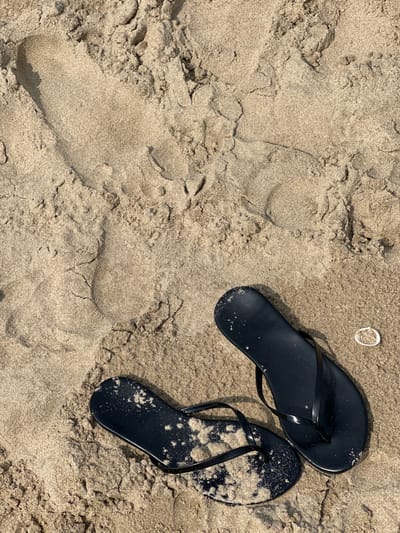 This is an overhead, eye-level shot of black flip-flops resting on a sandy beach. The sand is light brown and textured with ripples and small indentations, suggesting it's been disturbed by wind or water. Two distinct footprints are visible in the sand to the left of the flip-flops, with one print being deeper and more defined than the other. A small, white shell is situated to the upper right of the flip-flops. The flip-flops themselves appear to be made of rubber or a similar material, and one of them is dusted with a fine layer of sand. The lighting suggests it's daytime, likely sunny due to the shadows cast by the flip-flops and the shell, but the weather is not explicitly determinable beyond that. The scene evokes a sense of leisure and relaxation, typical of a beach setting. There is no visible text in the image.