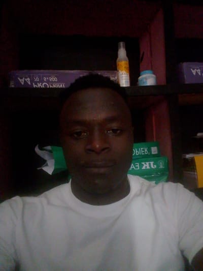 The image is a selfie taken from the chest up of a person. The person is wearing a white t-shirt and has dark skin. They are looking directly at the camera. In the background, there are shelves with various items, including a bottle of spray, a container, and stacks of paper. The lighting is dim, suggesting the photo was taken indoors in low light conditions. The specific location in the city of Mbar, Nigeria, cannot be determined from the provided image.