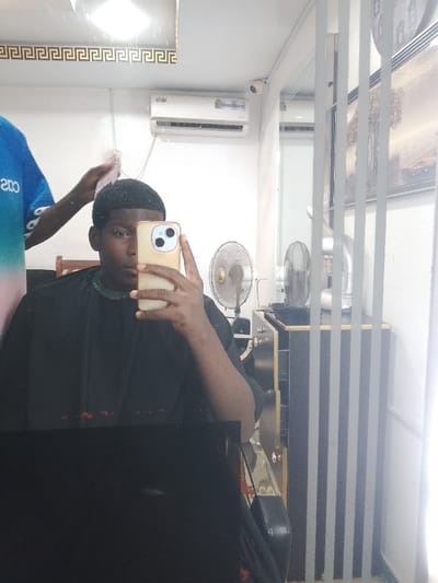 This image is a mirror selfie taken inside a barbershop or salon located in Mgbuoshimini, Nigeria. The primary subject is a young Black man, seated in a barber's chair and covered by a black cape, holding a light gold-colored smartphone to take the photo. The phone obscures part of his face. He has short, dark, curly hair.

Behind him, and also visible in the mirror's reflection, is another individual, presumably the barber, whose hands are near the main subject's head, indicating a haircut is in progress. The barber is wearing a blue and pink gradient shirt, with the partial text "Coo" visible on it.

The background, reflected in the mirror, shows typical barbershop elements. Above the main subject, a white LG air conditioner unit is mounted on the wall, alongside a small CCTV camera. The top edge of the mirror features a decorative gold border with a Greek key pattern. To the right, two standing fans are visible, along with a dark cabinet or dresser and a framed picture or artwork. The setting appears to be indoors during the daytime. The black barber's cape has subtle red markings or patterns along its lower edge.