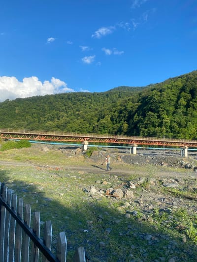The image captures a scenic view in Yuxarı Tala, Azerbaijan. A steep, tree-covered mountain dominates the left side of the frame, its lush green foliage contrasting with the clear blue sky dotted with scattered white clouds. A bright, sunny day is indicated by the strong sunlight casting shadows.
Running along the base of the mountain is a sturdy metal structure, likely a pipeline or aqueduct, supported by concrete pillars. A person, appearing to be a lone male wearing a light-colored shirt and dark pants, stands on a dirt path next to the structure, looking towards the mountain. The path is rocky and appears to be a utilitarian route, possibly for maintenance of the pipeline.
To the right of the path, the terrain is rough, with a mix of rocks and sparse green grass. In the foreground on the far right, a set of wooden stairs with a metal handrail leads down, suggesting a point of access or observation. The overall impression is one of a natural landscape integrated with industrial infrastructure, likely in a rural or mountainous region. The time of day appears to be mid-morning or afternoon, given the angle of the sun. There is no visible text in the image.