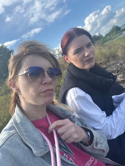 In an outdoor scene near Shuiskaya Chupa, Russia, two women are captured in a selfie. The woman in the foreground, positioned on the left, has light brown hair and is wearing large, reflective sunglasses and a light blue denim jacket over a bright pink sweatshirt. Her hand is visible, holding onto a pink drawstring. The second woman is on the right, with reddish-brown hair, wearing a black puffer vest over a white shirt. Both women are looking towards the camera, with the woman on the right appearing to be looking slightly away. The background consists of dry grass, rocky ground, and a hazy sky with scattered clouds, suggesting a natural, open landscape.