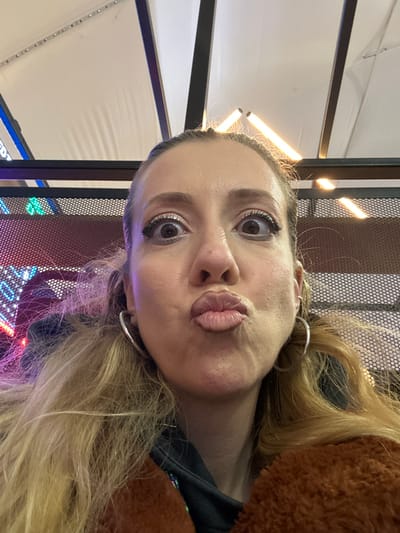 A close-up selfie of a woman with light skin and blonde hair. She is wearing silver hoop earrings and a brown jacket. Her expression is a pout. Behind her is a mesh-like surface with LED lights and a black metal frame. Overhead is a white surface with linear lighting fixtures. The location appears to be an indoor arcade or entertainment center, judging by the lights and game-like environment. The exact location in Dartford, United Kingdom cannot be determined from the image alone.