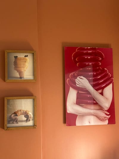 The scene depicts an interior wall space, described as being in Acassuso, Argentina, painted a uniform terracotta or orange-brown color. An internal corner is visible in the upper-left quadrant. Three distinct artworks are displayed on the wall.
To the right of the corner, a large, rectangular, vertically oriented print or poster is mounted. This artwork features a bare-chested individual, appearing female, shown from the mid-torso upwards against a solid red background. The individual's head and neck are entirely obscured by multiple concentric, glossy, dark red, ring-shaped tubular structures stacked vertically. Additional similar rings wrap around the individual's upper chest and shoulders. The person's arms are visible, with hands placed on the chest and lower abdomen, seemingly interacting with the red structures.
To the left of the corner, two smaller, square-framed artworks are stacked vertically. Both frames are light brown wood. The upper artwork shows a light-colored, vertically oriented, woven or spiral-shaped structure, resembling a vase or beehive, with a narrower base and an irregular, wider top. Two small, dark, mouse-like figures are perched on the upper rim of this structure. The background of this print is a muted light gray. The lower artwork depicts a naked human figure in a quadrupedal stance, arching its back. A series of light-colored, tubular, ring-shaped objects forms an arch over the figure's back. Another partially visible human form, possibly a second figure or a continuation of the first, is positioned to the right of the main figure. The background of this print is also a muted light gray.