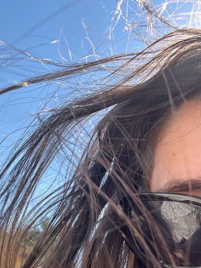The image is a close-up shot of a person's face and dark brown hair, which is blowing in the wind against a bright blue sky. The person is wearing dark sunglasses. The lower right portion of the image shows the upper part of a cheek and an eyebrow. The scene appears to be outdoors, possibly in Νέα Ηρακλείτσα, Greece, given the clear blue sky. The sunlight is catching the strands of hair, creating a glinting effect.