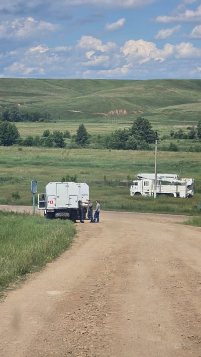 In Ekaterinovka, Russia, on a dirt road bordered by tall green grass, three men in work clothes stand near a white utility truck with its rear door open. One man leans against the truck, while another gestures with his hand, and the third stands with his arms crossed. A second white truck with a large crane is parked further down the road, partially in a grassy field. In the background, rolling green hills meet a cloudy blue sky.