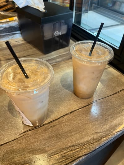 The image shows two clear plastic cups filled with a light brown beverage, likely iced coffee or a similar drink, each with a black straw. They are sitting on a wooden-like surface that appears to be a table or counter. Next to one of the cups is a black tissue dispenser with some tissues visible. The location appears to be a cafe or coffee shop in Fredericksburg, USA, based on the generic nature of the scene and assuming the photographer is located in Fredericksburg. There are no people visible in the image. The dominant action is the presence of prepared beverages ready for consumption.