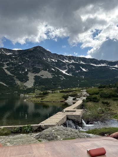The image depicts a scenic mountain lake with a dam structure. The dam, constructed of concrete blocks, runs across a narrow channel where water flows, creating a small waterfall. The lake's surface is calm and reflects the surrounding landscape. On the far side of the lake, a lush green mountainside rises, dotted with trees and patches of snow, culminating in a rocky summit. The sky is partly cloudy with dramatic formations of white and grey clouds against a blue backdrop. In the foreground, a wooden surface, possibly a table, is visible, with a red object and a hand holding a red object resting on it. The scene suggests an outdoor recreational area. The specific location is Avramovo, Bulgaria.