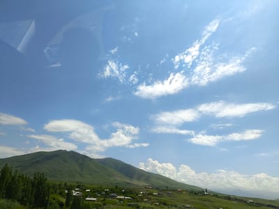 The image shows a landscape view of Artashavan, Armenia, under a bright blue sky with scattered white clouds. A large, green, rolling mountain dominates the background, with its slopes covered in lush vegetation. In the foreground, a small village is nestled among trees and fields. The village consists of several houses with visible rooftops, some appearing to be made of light-colored material, possibly solar panels. Tall, slender green trees line the left side of the frame, partially obscuring the view of the village and the mountain. The overall scene depicts a rural setting with a clear, sunny day. A reflection of what appears to be a car interior, including a side mirror and window frame, is visible in the upper left quadrant of the image, suggesting the photograph was taken from inside a vehicle.