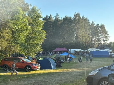 The image captures an outdoor gathering, possibly a festival or event, in the area of Valozhyn in Belarus. The scene is set in a grassy field with a backdrop of dense pine trees under a clear blue sky. In the foreground, an orange station wagon is parked, with a young person in athletic wear running past it. Several tents are pitched across the field, in various colors including blue, green, and purple. In the mid-ground, a group of people are seen gathered, some standing and others near motorcycles. Further back, larger tents or canopies are visible, suggesting vendor stalls or larger gathering areas. The overall atmosphere appears relaxed and communal, with the sun casting long shadows, indicating it might be late afternoon or early evening.