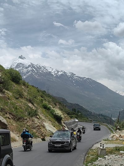 An overcast, cloudy sky looms over a winding mountain road in India. In the foreground, a black Kia Seltos SUV with license plate HR05BK9338 is driving on the asphalt road, with its front passenger visible. To its left, partially visible, is the rear of a dark-colored Jeep. Behind the Kia, several motorcycles are riding in a convoy. The first is a black motorcycle with a rider wearing a blue jacket, followed by two more motorcycles, one red and one black, also with riders. The road curves to the right, flanked by steep, rocky slopes covered in green vegetation and some sparse white flowering bushes. In the distance, imposing snow-capped mountains dominate the landscape, their peaks sharp against the cloudy sky. A power line pole is visible on the mountainside. The overall scene depicts a journey through a mountainous region.