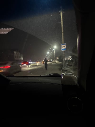 View from inside a car at night. A poorly lit street is visible through the windshield, with a person standing in the middle of the road, appearing to be wearing dark clothing. A blue and white bus stop sign stands on a pole to the left. Streetlights illuminate the scene, with several vehicles with red tail lights visible further down the road. The interior of the car is dark, with only a portion of the dashboard and windshield visible.