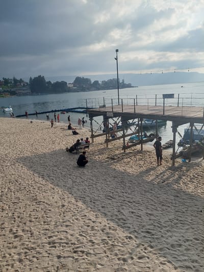 The image shows a sandy beach area on the shore of Lake Toba in Parapat, Indonesia. A wooden pier extends over the water, with several boats docked underneath and nearby. People are scattered on the beach and in the shallow water. Some individuals are sitting on benches, while others are walking or standing. In the background, there is a line of buildings and lush green hills under a cloudy sky. The lighting suggests it may be late afternoon.
