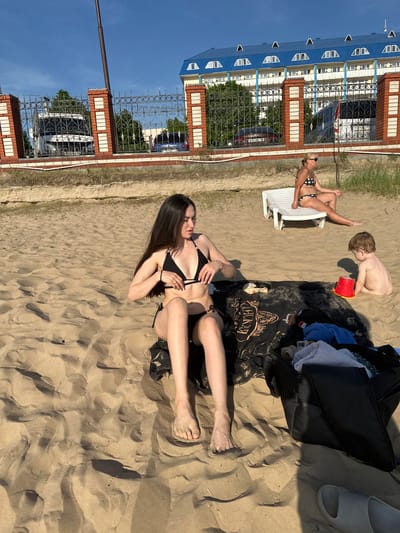 The image depicts a beach scene in Matiushino, Russia. A young woman with long dark hair, wearing a black bikini, is sitting on a dark-colored beach towel on the sand, adjusting her bikini top. Beside her is a black beach bag, containing various items including towels and a pair of white sandals. A young boy is partially buried in the sand, holding a red plastic bucket. Nearby, an older woman in a polka-dot bikini is sitting on a white plastic beach chair. In the background, there's a fence made of brick and wrought iron, behind which are buildings with blue roofs and white walls. The sky is clear and blue.